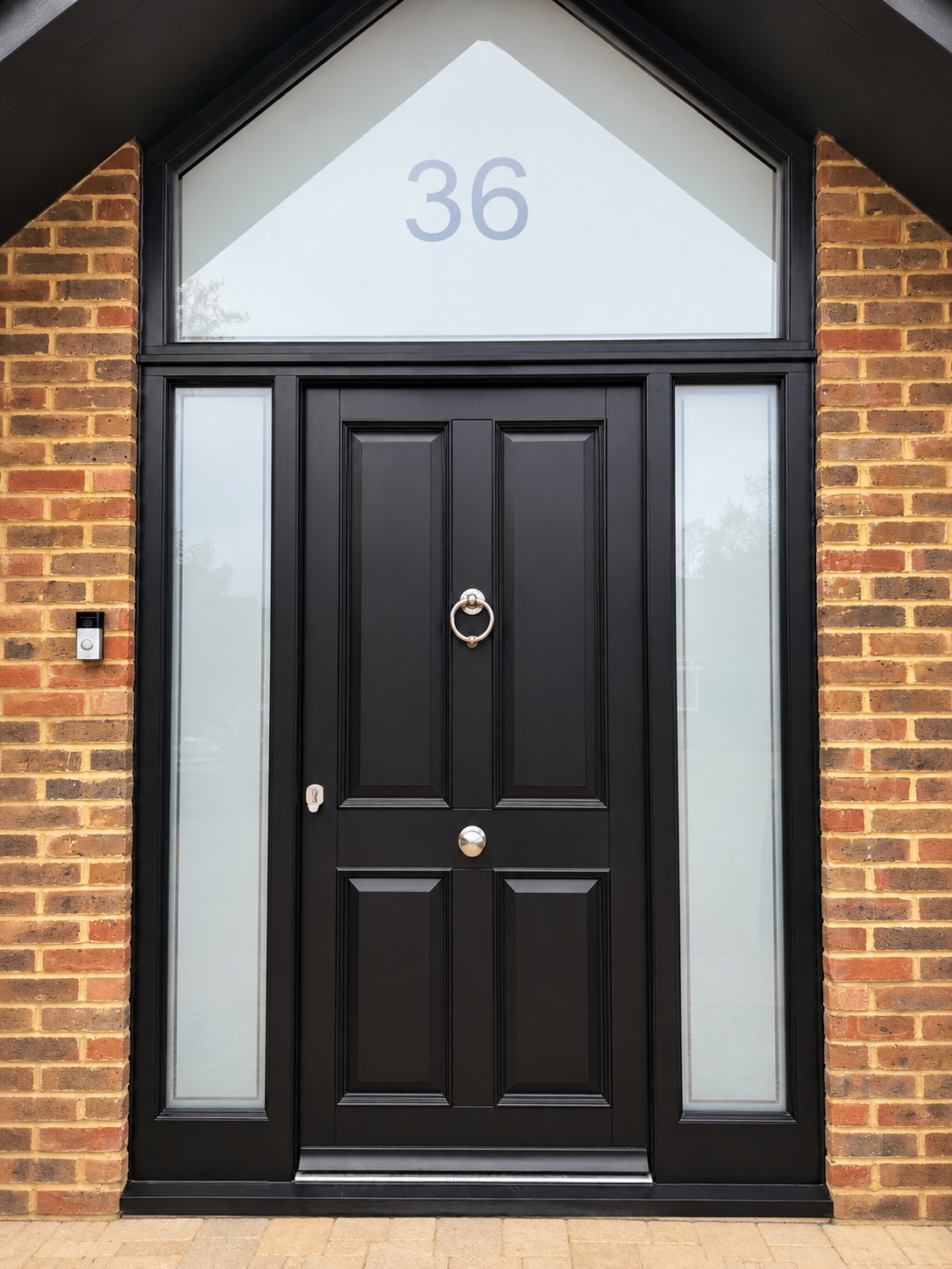 Black painted timber front door with glazed side lights and an apex gable window above in a brick porch